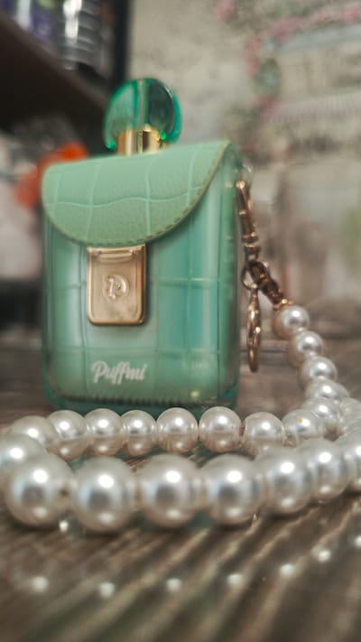 A green, purse-shaped perfume bottle with a textured lid sits on a wooden surface, partially obscured by a strand of pearl beads in the foreground. The bottle has a gold clasp with a "P" logo and the word "Puffmi" printed below it. A gold chain is attached to the side of the bottle, with a small charm hanging from it, also partially covered by pearls. The background is blurred and appears to be an interior setting with shelves and objects. There is no indication that this scene is located in Zvenigorod, Russia.