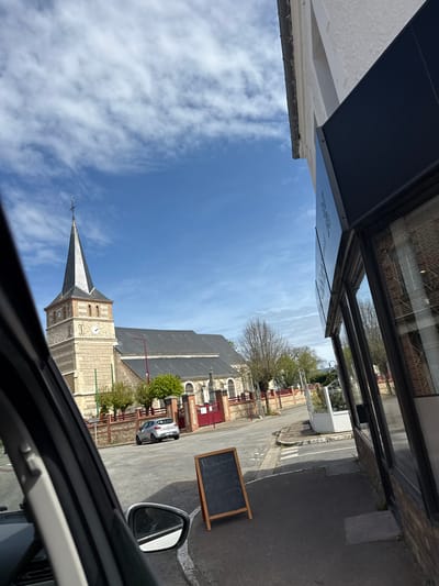 This media file captures a sunny daytime street scene in what appears to be a small town or village, viewed from inside a vehicle looking out a side window. The overall impression is one of a quiet, rural European setting, consistent with Le Tilleul, France.
The most prominent subject is a stone church with a tall, grey spire dominating the mid-ground. Its tower features two visible clock faces. Surrounding buildings, some with red-painted window frames or shutters, line the street. A bright blue sky with scattered white, wispy clouds fills a large portion of the frame.
In the foreground, the structure of the vehicle's interior, including a side mirror, frames the view. To the left, a dark-colored awning of a building is visible, with some ornate, reflective text that is largely illegible. Below this, a white building facade with a dark gutter is seen.
The asphalt road shows painted markings and a crosswalk. A silver hatchback car is parked on the street in the distance. On the sidewalk, a black chalkboard sign stands, displaying visible text that reads "Restaurant" at the top, followed by "OUVERT" (French for "OPEN") and other indistinct menu items or specials below. Bare trees suggest either early spring or late autumn. No people or overt activity are visible in the static scene. The lighting indicates a bright, clear day.