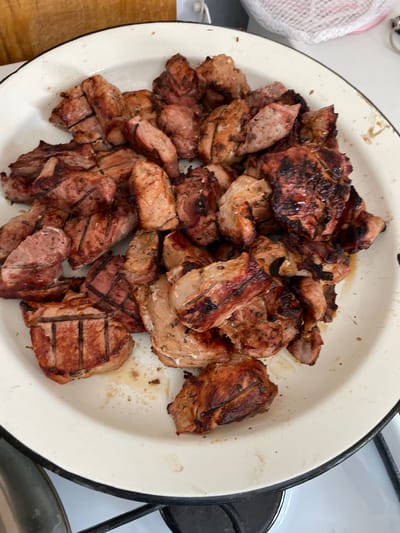 A top-down shot shows a white enamel plate filled with grilled meat pieces, seemingly on a stovetop, possibly in Zapadnaya, Russia. The meat pieces vary in size and shape, displaying grill marks. The plate is old with a black rim and some food stains. The background includes sections of the stove and a wooden surface. No people are visible.