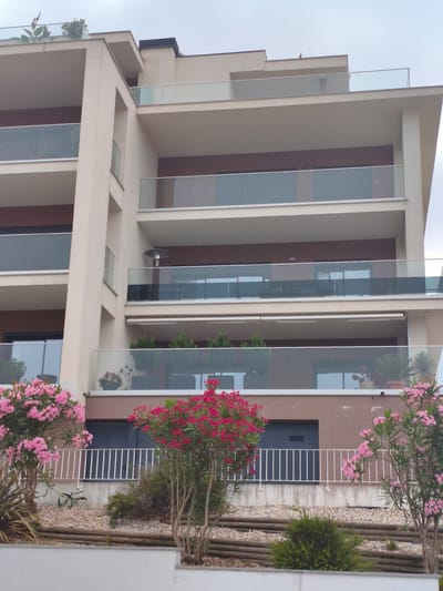 The image shows a modern, multi-story building with balconies. The building is a light beige color and has dark brown accents. The balconies are enclosed with glass railings. There are several trees and bushes in front of the building, including some with pink flowers and others with red flowers. The sky is overcast. There are no people visible in the image. The location is identified as Nazaré, Portugal.