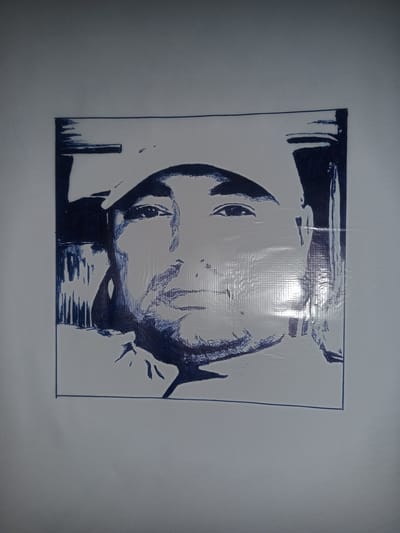 This is a close-up, black and white, ink drawing of a man wearing a baseball cap. The drawing is framed within a black border. The man is depicted from the chest up, facing forward. His eyes are looking directly at the viewer. He has a mustache and a slight smile. The drawing utilizes hatching and shading techniques to create depth and form. The background is white, with some abstract blue lines suggesting an environment. There is a slight glare on the surface of the drawing, indicating it might be a photograph of a physical drawing. The overall mood of the drawing is neutral and direct. The drawing is situated within a location context of Gulbag, Uzbekistan, but there are no explicit visual cues in the image to confirm this location. No text is visible in the image.