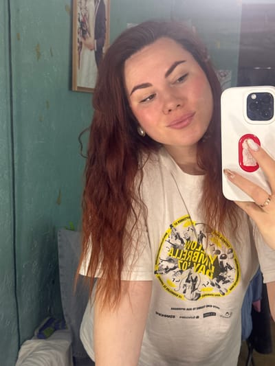 A woman is taking a selfie, holding a white iPhone with a red sticker on the back. She has auburn hair, fair skin, and dark eyebrows. She's wearing a white t-shirt with a yellow and black graphic design. A picture frame hangs on a green wall, likely in Черёмушки, Russia. The background shows a glimpse of casual clothing. The text on the t-shirt includes the words "TO," "COM," "CHIZMICK," and "MAF LETTOALETYPECON." Other text is reflected, reversed and illegible.
