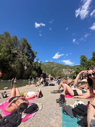 The image shows a crowded riverbank scene in Montanejos, Spain. The river is lined with people sunbathing and swimming. Most people wear swimwear or casual clothing. The river is green, and there are trees and a rocky hill on the other side. Some people are lying on towels on the riverbank. The sky is blue with scattered clouds. The riverbank is made of gravel. Some people are wading or swimming in the river.