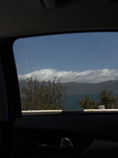 The image shows a view from inside a vehicle, looking out through a window at a scenic landscape. The foreground of the image is dominated by the interior of the car, with a dark colored door panel and a door handle visible. Above the door panel, a light-colored horizontal bar runs across the bottom of the window.
The view through the window reveals a body of water, likely a lake, with the surface rippling under a bright sky. Beyond the water, rolling hills or mountains are visible, covered in a muted green or grey vegetation. A band of fluffy white clouds stretches horizontally across the sky, partially obscuring the tops of the hills. In the mid-ground, a cluster of trees with green foliage adds depth to the scene. The lighting suggests it is daytime, with the sun likely positioned overhead or slightly to the side.
The location is identified as Tsamakaberd, Armenia.