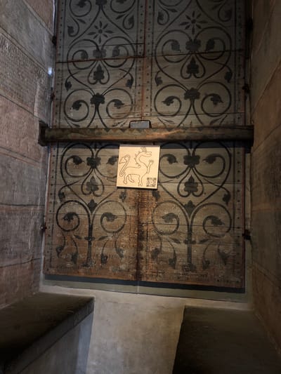 The image depicts a section of the Chillon Castle in Veytaux, Switzerland. Two large, aged wooden doors are visible, each decorated with a symmetrical, dark gray floral and vine pattern. A central vertical beam, also patterned in dark gray, separates the two doors. A light brown sign with a black cartoon drawing and a QR code is affixed to the right door. The floor is made of textured gray stone tiles with thin darker gray lines running across. The background includes parts of a concrete wall with a built-in ledge on the right side. The scene suggests an interior passage within the castle.