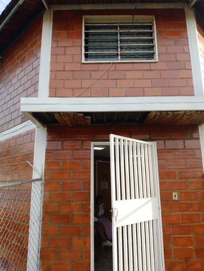 A two-story brick building with white trim stands in Barcelona, Venezuela. The building has a window with horizontal slats on the upper story and an open white metal gate on the lower story. A wire runs along the brick wall to the window. A woman sits inside the building behind the open gate. A chain-link fence adjoins the building on the left side. The environment is brightly lit.