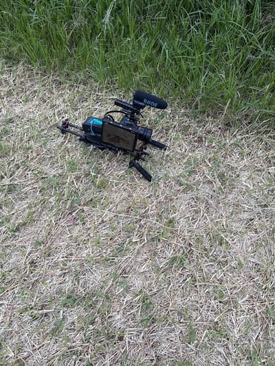 A professional video camera rig is positioned on a grassy surface in El Paraíso, Mexico. The rig consists of a dark-colored camera body equipped with an external monitor displaying an image, a 'RODE' brand shotgun microphone with a black foam windscreen mounted on top, a visible battery pack, and two integrated handles. The entire assembly rests on a small, black, collapsible tripod. The immediate ground cover is composed of dry, light brown grass interspersed with green blades, transitioning into a denser patch of vibrant green grass in the background. No individuals are present in the scene, and no actions or interactions are depicted.