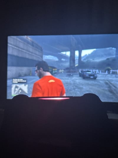 The image displays a digital screen captured in a dimly lit environment, showing a scene from the video game Grand Theft Auto V (GTA V). The lower portion of the screen is obscured by the top of a dark-colored game controller, which features a central light bar emitting a reddish-pink illumination.
The game scene itself portrays a male character, viewed from the back, slightly turned to his left. He has dark hair, wears glasses, and is dressed in a solid orange t-shirt with a small, yellow, indistinct logo on the upper back. The character stands on an irregular, unpaved ground surface, which appears to be dirt or gravel. To the left, a large, unadorned grey concrete wall or building rises. In the background, elevated concrete highway structures or an overpass are visible against a bright, hazy sky, indicating daylight. A dark-colored sedan is stationary in the mid-ground to the character's right. Distant hills or mountainous terrain are discernible beneath the elevated structures in the far background. A mini-map and other unidentifiable user interface elements are visible in the lower-left corner of the screen.
The screen and game controller are physically located in Fowlis Easter, United Kingdom.