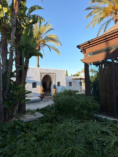 The image depicts a sunlit courtyard with lush vegetation and a white building. A wooden overhang with intricate carvings shades a portion of the scene. Palm trees and broad-leafed plants frame the view, their fronds reaching towards a clear blue sky. The white building features a distinctive arched entryway decorated with detailed plasterwork and two arched windows with ornate grills. The architecture suggests a Mediterranean or North African influence, consistent with the location in Sant Joan de Labritja, Spain. A patio umbrella is partially visible near the building. The lighting indicates it is daytime, with bright sunlight casting shadows. The overall atmosphere is tranquil and inviting, evoking a sense of peace and warmth. No people are visible in the frame.