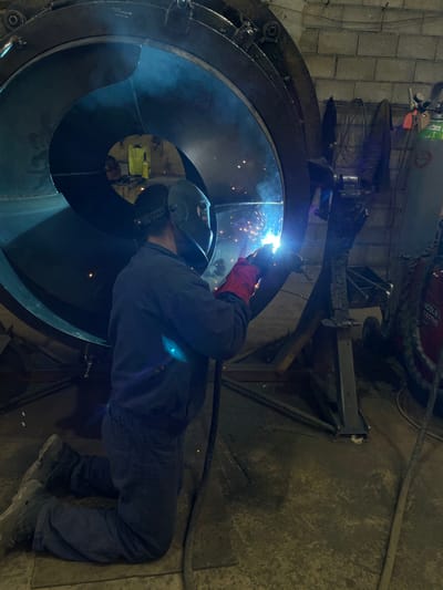 A person is welding inside a large, circular metal structure. The welder wears protective gear including a helmet and gloves. Sparks and smoke are visible from the welding process. The environment appears to be an industrial workshop, based on the equipment and materials in the background. The location of Tomba, Montenegro, is not evident from the image.