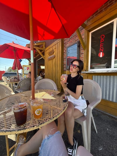 The image depicts an outdoor seating area of a restaurant, possibly located in Hampton, United States of America. A young woman with dark hair, wearing sunglasses, a black t-shirt, and white shorts with black stripes, sits in a chair. She is holding a pink beverage with a straw and appears to be pouting or making a face. On a yellow metal table in front of her are two clear plastic cups with straws, one containing a dark liquid, possibly iced coffee, and the other a lighter-colored beverage with a "Darling's Bee" sticker. A smartphone and a cigarette pack are also visible on the table. In the background, a building with wooden siding and a brick section is visible. A red umbrella provides shade over the seating area. The overall impression is of a casual outdoor dining experience on a sunny day.