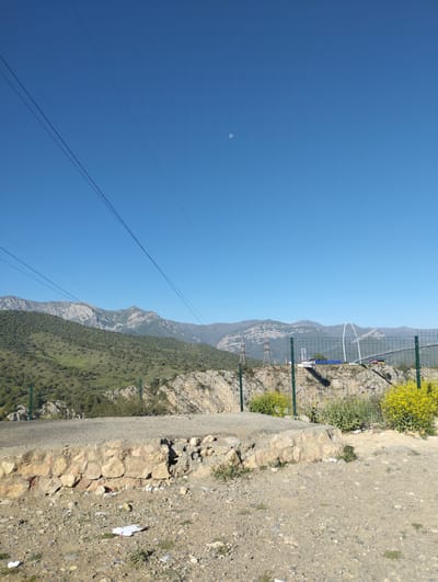 A wide shot captures a clear, bright blue sky with a faint moon visible in the upper center of the frame. Three power lines stretch diagonally from the top left corner towards the center. In the middle ground, a majestic mountain range dominates the landscape, with slopes covered in lush green trees and dotted with rocky outcrops. A few electrical towers and a very long, arched bridge are visible in the distance, along with a sign that reads "Pedal Boats." The foreground consists of a dirt path with scattered rocks, some wildflowers, and a low stone wall. A few pieces of white paper are scattered on the ground in the foreground. The image appears to be taken from a vantage point overlooking a valley with a distant bridge. The location is identified as being in the city of Myk, Uzbekistan.