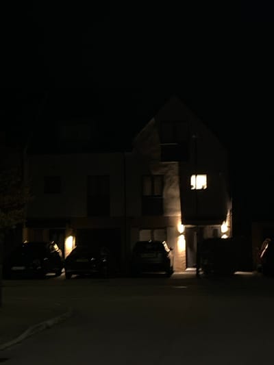 The image displays a nighttime scene in a residential area of Stevenage, United Kingdom. A modern, light-colored, three-story building with a pitched roof section on the right occupies the majority of the frame. The scene is predominantly dark, with artificial illumination provided by several sources. A single upper-story window on the right side of the building is brightly lit from within, casting a rectangular glow. Multiple wall-mounted up-and-down lights illuminate the building's facade, particularly near the ground level and along the central section. Three dark-colored passenger vehicles are parked in front of the building on a paved surface, which appears to be a driveway or parking area. A distinct curb line is visible in the lower foreground. The sky is dark, and minimal foliage is discernible on the far left. No individuals, actions, or interactions are present in the static scene.