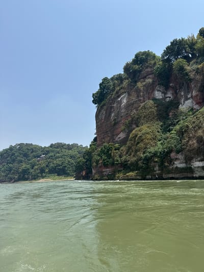 The image captures a scene in Shizhong, China, featuring a river adjacent to a steep cliff face. The river water is a murky green color. The cliff face is primarily composed of rock formations, with patches of green vegetation clinging to its surface. The sky above is a pale blue. The overall perspective suggests the photo was taken from a position on the river.