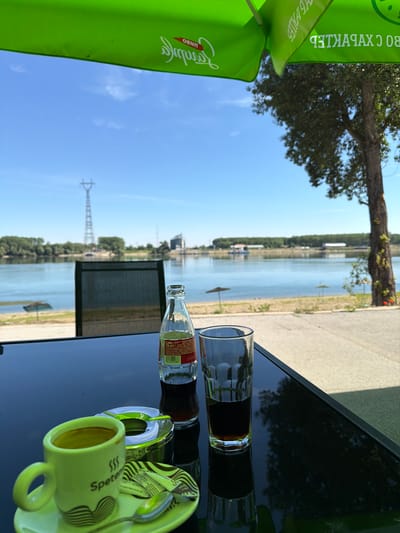 A close-up shot features a glass and a glass bottle of Coca-Cola on a glossy black table. The bottle is partially obscured by the glass, and its label is visible with text that includes "Coca-Cola" and "250 ml." To the right, a pale green ceramic cup sits on a matching saucer, with a spoon resting on the saucer. The cup has a wavy line design and some text in a dark color, including what appears to be "Spetema."

In the background, a wide river is visible under a bright blue sky with scattered white clouds. Trees line the riverbank, and a power pylon stands in the distance. To the left of the frame, a large section of a bright green umbrella or canopy is visible, with some text printed on it in white, including "BO C XHAX9IKLEB" and possibly "VEG PALES." The lighting suggests it is daytime, likely sunny, given the clear sky and shadows on the table. The overall scene evokes a relaxed outdoor setting, possibly a cafe or restaurant by a river. The orientation of the image is vertical. The location is Oryahovo, Bulgaria.