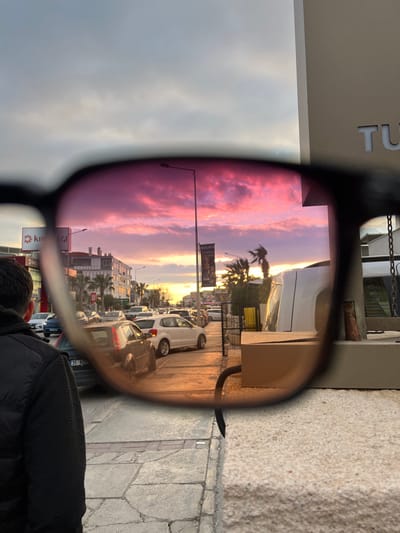 The image displays an urban street scene at sunset in Güzelbahçe, Turkey, viewed prominently through the dark-framed, rose-tinted lenses of sunglasses. Through the lenses, a vivid sunset dominates the sky, featuring clouds illuminated in shades of purple, pink, and orange. Below this horizon, a multi-lane road is visible, containing several parked and active vehicles, including a white Volkswagen Polo and a dark grey Fiat Punto (license plate "35 KEG"). Multi-story buildings line the street, one prominently displaying a red "KIRÇİÇEĞİ" sign. Silhouetted palm trees are visible against the colorful sky, alongside streetlights and a banner advertising "MİA LİFE Online."

Outside the left lens, the back of an individual's head and left shoulder, clad in a dark quilted jacket, is present. To the right of the lenses, a light-colored concrete barrier or wall forms the foreground, from which a black corrugated pipe extends horizontally towards a light brown cardboard box. Part of a vertical metal structure, from which a chain hangs, is partially visible behind the box. The sky observed outside the sunglass lenses is overcast and grey, contrasting with the filtered view. The immediate ground consists of a paved sidewalk composed of grey and reddish-brown square tiles.