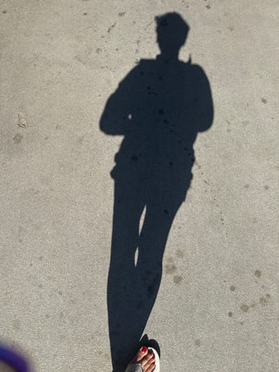 A shadow of a person standing with their arms crossed is cast on a grey, speckled concrete surface. The shadow is elongated and appears to be cast by a person wearing a hat and possibly a jacket. The person's feet are visible at the bottom right of the image, wearing open-toed sandals. The scene appears to be outdoors, likely on a sidewalk or paved area.