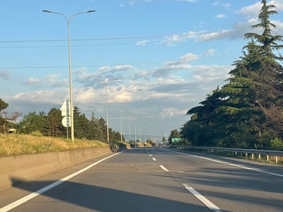 The image depicts a highway scene in Akhalsheni, Georgia. The perspective is from a vehicle driving on the road, looking straight ahead. The highway has multiple lanes, separated by white lines. On the left side of the road, there is a concrete barrier, followed by a grassy embankment and a line of trees and bushes. On the right side, there's a metal guardrail and a dense line of trees, including a prominent evergreen tree. Several streetlights line the highway, illuminating the road. The sky is blue with scattered white clouds, suggesting it's daytime, likely late afternoon or early evening due to the warm lighting and shadows. The overall atmosphere is calm and serene, with no people visible. There is a green road sign in the distance on the right side of the road, with text in what appears to be Georgian script. The overall impression is of a typical highway journey through a green, somewhat rural landscape under a pleasant sky.