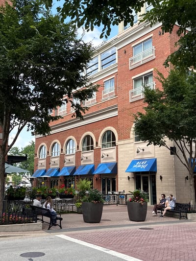 In the city of Naperville, United States of America, a brick building with multiple arched windows and blue awnings stands adjacent to a paved walkway. Two couples are seated on benches outside the building, and several large planters with flowers are placed on the walkway. Trees with green foliage are visible in the foreground and background. The sky is partly cloudy.