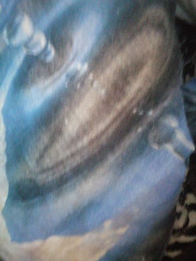 The image is a close-up, abstract photograph of a fabric with a printed pattern. The pattern appears to be a stylized representation of a nebula or galaxy, with swirling blues, grays, and browns. There are no discernible people, objects, or actions within the image. The environment is therefore not applicable, and no location in Milzkalne, Latvia, can be identified.