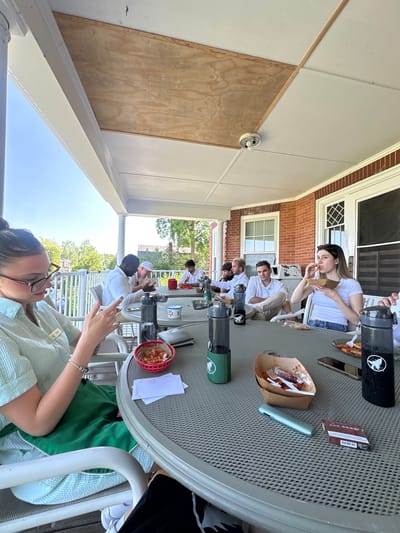 A group of eight people are gathered around a round table on an outdoor porch, eating and socializing. The porch is covered by a wooden ceiling with exposed beams. The individuals are dressed casually, and most are looking at their phones or food. Food containers, bottles, and napkins are scattered on the table. The background shows trees and a brick building. The scene suggests a casual outdoor dining experience.