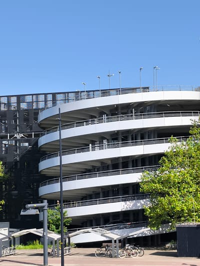 The image depicts a multi-story parking structure in Hoofddorp, Netherlands, characterized by a spiral ramp on the right side. The building is primarily white with gray accents. The structure is partially obscured by a tree with green leaves in the foreground. Adjacent to the parking structure is a black metallic grid structure. A bike rack shelters several bicycles in the foreground. The sky is a clear, bright blue.