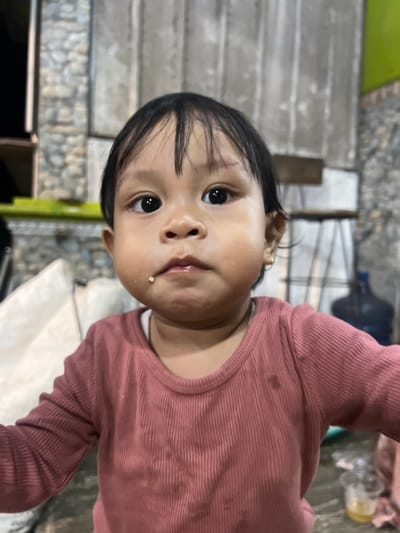 A close-up, sideways shot features a young child with dark hair and dark eyes, looking directly at the camera. The child is wearing a pink, ribbed long-sleeved shirt. A small piece of food is visible on their lower lip. The background is blurry, but appears to be an outdoor setting with concrete steps and some greenery. The lighting suggests it is daytime. The image is candid and intimate, capturing a simple moment with the child. The location context provided is Wedarijaksa, Indonesia.