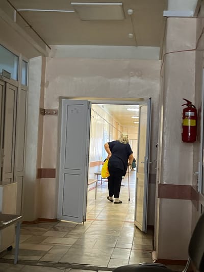 The image depicts an indoor hallway, likely in a medical facility, with a woman using a cane walking away from the camera and towards an open doorway. The hallway has tiled floors and beige walls, with a red fire extinguisher mounted on the right wall. Fluorescent lighting is visible on the ceiling. The woman is carrying a yellow bag and is dressed in dark clothing. The scene appears to be an ordinary day in a public space.