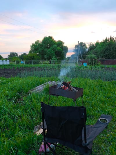 A black folding chair is positioned in the foreground, angled towards a metal brazier containing burning wood and emitting smoke. The brazier sits amidst tall green grass and other vegetation. Behind the brazier and chair, rows of plants are visible in a garden plot, with a fence demarcating the property line. Trees and a utility pole are present in the background, under a dusky sky with hints of pink and orange. The scene appears to be a rural or suburban outdoor setting in Yaganovka, Russia.