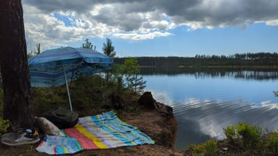 The image depicts a serene lakeside scene, likely in the vicinity of Severnaya Samarka, Russia, on a partly cloudy day. In the foreground, a large, blue and white striped beach umbrella is set up next to a thick tree trunk. Beneath the umbrella, a colorful striped blanket is spread out on the ground, accompanied by a green backpack, a patterned pillow, a pair of athletic shoes, and a partially obscured bottle of water. The terrain in the immediate foreground is earthy with some vegetation. The middle ground features the calm, reflective surface of a lake, with the opposite bank lined by a dense forest of pine trees. The sky above is a mix of blue sky and scattered clouds, some of which are casting shadows on the water. The overall impression is one of a quiet, natural setting prepared for relaxation.