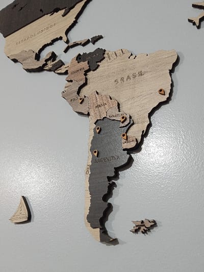 A wooden map of South America and parts of North America is mounted on a gray surface. The map sections representing the countries are crafted from varying shades of wood. Place markers indicate locations within countries such as Peru, Brazil, Paraguay, Uruguay, Chile, and Argentina. Two smaller wooden objects, a sailboat and the Falkland Islands, are positioned separately from the main map. The map includes country names etched into the wooden surfaces.