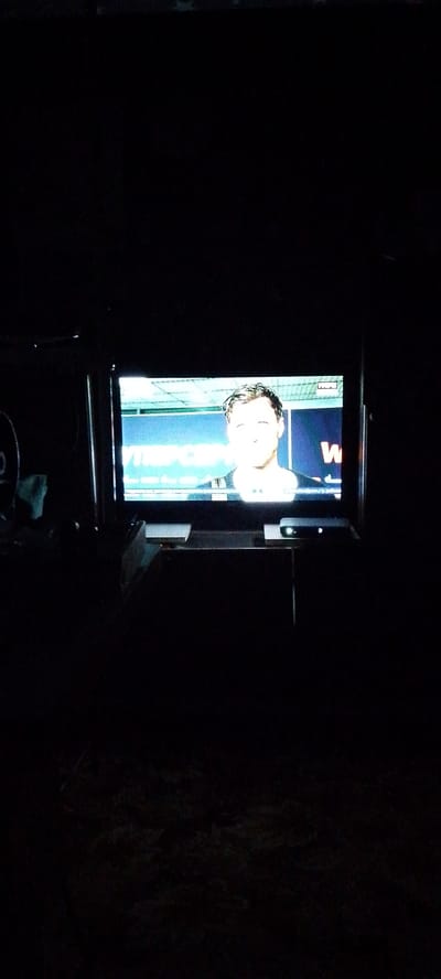 The image depicts a television screen displaying a close-up of a man's face. The man appears to be of Caucasian descent with short, light brown hair. He is wearing a dark shirt. Behind him, a blue backdrop is visible with some text, including the letter "W" in red. The television itself is dark, suggesting the surrounding environment is dimly lit or the photo was taken at night. A black electronic device with a small light on it is placed on top of a dark surface beneath the television. The overall scene is indistinct due to the low light conditions. The image does not provide any specific information that would allow for localization to Storozhi Pervye, Russia.