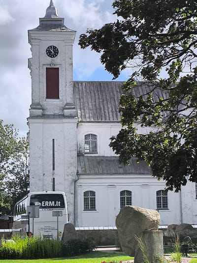 The image captures the exterior of a white church with a prominent clock tower, located in the town of Mosėdis, Lithuania. The clock on the tower shows the time as approximately 12:48. Adjacent to the church, a white bus with "ERM.lt" printed on its rear is parked. In the foreground, there is a grassy area with several large rocks, some of which are part of an art installation. A tree with dark green foliage partially obscures the view of the church's roof. The sky is overcast with some visible blue patches.