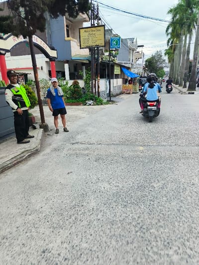 The image shows a street scene in Kecamatan Dayeuhkolot, Indonesia (latitude: -6.963, longitude: 107.596). A man in a black uniform and a yellow vest stands on the left side of the street, looking directly at the camera. He has a walkie-talkie on his belt. Another man wearing a blue shirt and a white hat is standing on the sidewalk to the right of the image, looking towards the street. Behind them, a building with a sign that reads "Aisha Skin Care Salon" is visible. A motorbike with two passengers is driving towards the camera on the street. In the distance, there are more buildings and trees along the street. The sky is cloudy.