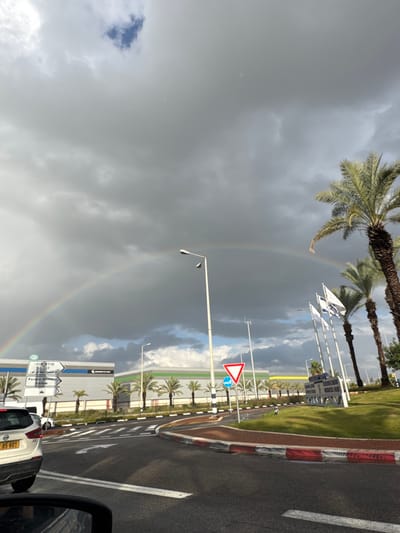 The image depicts an outdoor scene at what appears to be a road junction or roundabout in Pardes Hana-Karkur, Israel, under a heavily clouded sky with a visible rainbow. The viewpoint is from inside a vehicle, evidenced by a partial rearview mirror in the lower left and the rear of a white car with an Israeli license plate (e.g., 85-801) in the left foreground.

The sky is predominantly covered by dark, stormy gray clouds, with a segment of blue sky visible towards the upper left. A distinct rainbow forms a broad arch across the mid-upper section of the sky, spanning from left to right.

In the midground, a multi-lane asphalt road is visible with white lane markings. A red and white painted curb borders the road on the right. A prominent white pole with a street lamp stands centrally. To the right, a red-bordered white triangular "Give Way" (yield) sign is present, along with a blue circular sign. A grassy median or island features several tall palm trees. Further right, a cluster of flagpoles bears what appear to be Israeli flags. A large white directional road sign, displaying Hebrew and English text including "כביש 653" (Road 653) and "Pardes Hana-Karkur," is visible on the left side of the road.

In the background, behind the road and palm trees, a row of large, modern industrial or commercial buildings with horizontal grey/blue and green/yellow striping on their facades is visible. More palm trees are situated around these buildings. No individuals are explicitly visible. The scene suggests a recent or ongoing period of rain, given the clouds and rainbow.