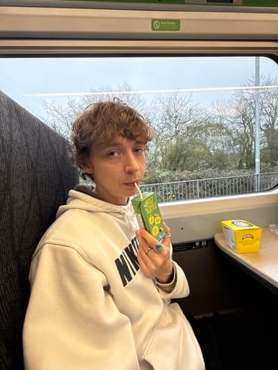 A person with short, curly brown hair and a visible nose piercing is seated inside a Great Western Railway train carriage, traversing the district of Mid Devon, United Kingdom. The individual, wearing a light-colored hoodie with a "NIKE" logo and two rings (one plain silver, one with a turquoise stone) on their left hand, holds a green "MEET THE WONKY SQUAD" juice box with a straw to their mouth, looking directly at the camera. A yellow Marmite box rests on the adjacent table surface to their right. The train's interior includes grey upholstered seating, and an overhead panel features a "Quiet Carriage" sign. Through the window, a landscape of bare deciduous trees and shrubs is visible under an overcast sky, suggesting a cold season.