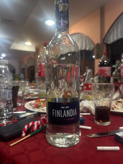 The image depicts an indoor scene in Arkovna, Bulgaria, specifically a dining table after or during a social gathering. The table is covered with a red tablecloth.

The central subject is a nearly empty, clear glass bottle of "Finlandia Vodka of Finland," positioned upright. To its left, a partially full clear plastic water bottle with a blue label is visible. Further left, a dark-colored smartphone or small electronic device rests on the tablecloth, beside a red, white, and green striped object resembling a candy cane on a stick.

To the right of the vodka bottle, a clear glass with a Coca-Cola logo contains a small amount of dark liquid. Scattered on the tablecloth in the foreground are several cigarettes, one partially unrolled, and two blue cigarette packs, one open. A silver dinner knife is also present.

Various white plates with remnants of food, consistent with salads or other dishes, are distributed across the table. Additional empty or partially full Coca-Cola bottles and other glasses are visible in the mid-ground and background. White napkins or paper towels are also present.

The background features light orange-hued walls, illuminated by circular overhead lights. Arch-shaped structural elements or doorways with white drapes are discernible in the distance. No individuals are visible in the frame; however, the arrangement of items suggests multiple participants at the gathering. The scene indicates recent consumption of food, alcoholic beverages, soft drinks, and tobacco.