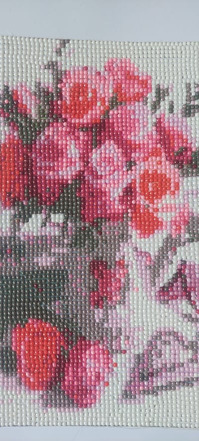 This is a close-up, vertical, eye-level shot of a diamond painting of flowers, rendered in shades of pink, red, and gray. The painting is composed of small, faceted resin beads. The composition is dense with floral elements, appearing abstract due to the extreme close-up. The background is a white border surrounding the artwork. The image does not contain any information about the city of Khobi, Georgia, or any other geographical location. There are no people, actions, or interactions depicted in the image.