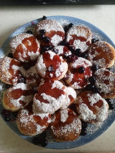 A close-up, overhead shot showcases a circular blue and white patterned plate filled with small, round, golden-brown pancakes, or possibly fritters. They are generously dusted with powdered sugar and drizzled with a thick, reddish-brown jam or syrup. Several dark blue blueberries are scattered amongst and on top of the pancakes, some appearing whole and others slightly crushed, releasing their juices. The plate rests on a light-colored, textured surface, possibly a countertop or table. The background is blurred, but appears to be a dark, reflective surface. No people are visible. The image does not provide any information about a specific location in Buraly, Russia.