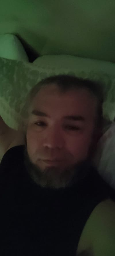This is a close-up, low-light, tilted selfie of a person lying down. The subject appears to be a man with short, dark hair and a beard. He is wearing a black tank top. His face is illuminated by a green light source, which casts a strong glow on the left side of the image and creates deep shadows on the right. The background is dark and out of focus, but suggests a soft surface, possibly a pillow or bed, on which the subject is resting. The overall mood is intimate and perhaps sleepy, given the low light and reclined posture. No text is visible in the image. The timestamp "2023-10-27 02:15" is overlaid on the image, indicating the photo was taken shortly after midnight.