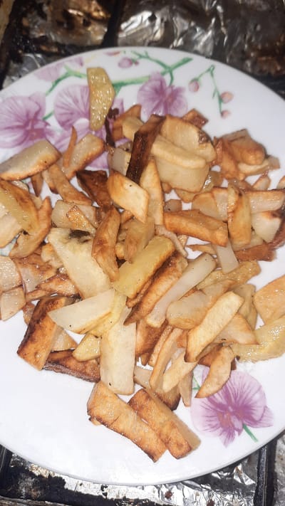 The image shows a close-up, overhead view of a white plate filled with a generous portion of french fries. The plate has a floral pattern with purple and pink flowers. The fries appear to be homemade, with varying shapes and sizes, some golden brown and crispy, others lighter in color. A few of the fries have a slightly darker, almost burnt appearance. The setting appears to be indoors, with a metallic, foil-like surface visible in the background, suggesting a cooking or baking sheet. There are no people visible in the image. The lighting is somewhat dim and appears to be artificial, casting a slight glare on some of the fries. The focus is entirely on the food, with no discernible text. The overall impression is a casual presentation of a popular comfort food.