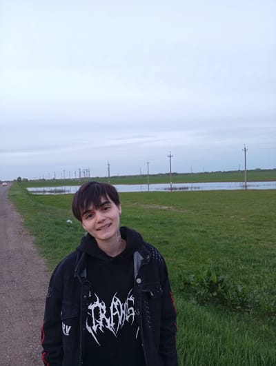 A young person stands on a dirt road in Azyakul', Russia, with a flooded field behind them. The person is smiling and wearing a black hooded sweatshirt with a white graphic design and a black denim jacket with red stripes on the sleeves. Power lines run along the field. A car is visible in the distance on the road. The sky is overcast.