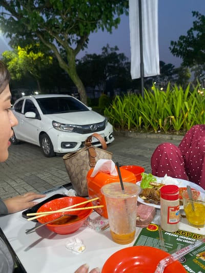This is a medium shot, eye-level, indoor/outdoor photograph taken at dusk in Mekar Bakti, Indonesia. The scene depicts a table with food and drinks set up in front of a white car parked on a paved surface. In the foreground, on the left, a woman's side profile is visible, with her looking to the left. She is wearing a grey top. To her right, on the table, are two red bowls with chopsticks and a spoon in one, a plastic cup with a beverage, and an orange container. To the right of the table, another person is partially visible, wearing a red top with white polka dots, with their arms resting on the table. In the background, there is lush green foliage and trees, with a white banner hanging from a pole. The overall setting appears to be an outdoor dining area.