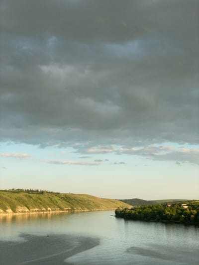 The image shows a landscape scene with a river or body of water flanked by land on both sides. One side features a grassy, elevated bank, while the opposite side is lined with dense trees and foliage. The sky above is partly cloudy, transitioning from a brighter, lighter blue towards the center to a darker, stormier gray on the left. The overall lighting is soft, suggesting either early morning or late afternoon.