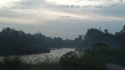 The image depicts a serene, hazy landscape of a river winding through dense vegetation. The sky is overcast with pale, elongated clouds, suggesting either early morning or late evening. The foliage on either side of the river is dark and indistinct, forming a silhouetted backdrop. The water itself appears slightly rippled and reflects the muted light of the sky. There are no people or man-made structures visible, emphasizing the natural and undisturbed environment. The overall atmosphere is tranquil and somewhat mysterious due to the soft focus and low light conditions. The scene is consistent with the natural riverine environments found in regions like Kubu, Indonesia, known for its lush tropical scenery and waterways.
