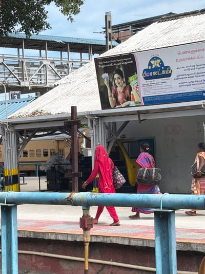 This is a photograph taken at a railway station in Thanjavur, India. The image is oriented vertically, showing a platform with people waiting. In the foreground, a person in a bright red outfit is walking away from the camera, their lower body and legs visible. They are wearing sandals. To the left of this person, a large blue metal pipe runs vertically. A sign for "Beemalakshmi" is prominently displayed, featuring a butterfly logo and text in Tamil. The sign advertises an insurance scheme for women, mentioning benefits like basic insurance coverage and additional sums. There's also a visual of a woman holding money, suggesting financial services.
Behind the people on the platform, a yellow structure, possibly a bench or play equipment, is visible. Further back, a train is partially seen. The setting appears to be outdoors during the day, with a cloudy sky overhead. The overall atmosphere seems to be that of a busy public space, with individuals going about their routines. The presence of the railway station, the train, and the Tamil text on the sign provide strong location cues. The people are dressed in traditional Indian attire, further reinforcing the regional context.