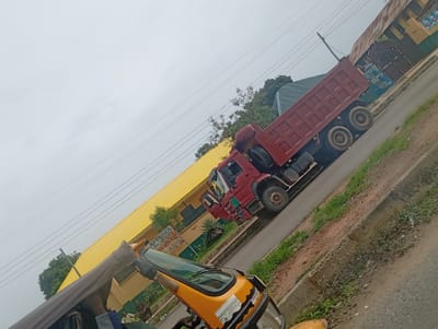 The image captures a street scene in Sabo, Nigeria. A red dump truck is parked on the right side of the road. To the left, a yellow auto-rickshaw (tuk-tuk) is also parked. In the background, there is a building with a yellow roof. A sign partially obscured by the auto-rickshaw reads "LAFENWA HIGH SCHOOL" and a road name, implying that it may be near Lafenwa Road. The sky is overcast. There are power lines overhead. The roadside has some patches of grass and dirt. The image appears to have been taken at a slight angle.