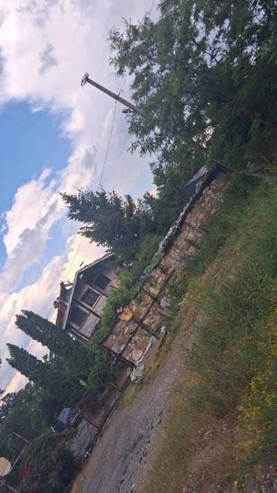 The image depicts a rural scene, likely in the village of Dobrotino, Bulgaria, characterized by a house with a stone foundation and weathered wooden siding. The house has multiple windows and a visible roof structure. Surrounding the house is dense vegetation, including trees and shrubs, with a gravel path leading towards it. A utility pole with wires is visible in the background, and a dark-colored vehicle is parked near a gate. The sky is partly cloudy, suggesting it is daytime. The overall impression is of a somewhat rustic and overgrown property.