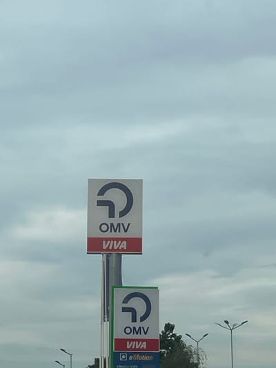 The image shows a cloudy sky over a street scene. Two OMV VIVA signs are prominently displayed on poles. The upper sign is larger and features the OMV logo, with "OMV" and "VIVA" text below. The lower sign, partially visible, has the same OMV logo and text, with additional text indicating "eMotion kWh". Streetlights and trees are visible in the background. This scene is located in Orizovo, Bulgaria.