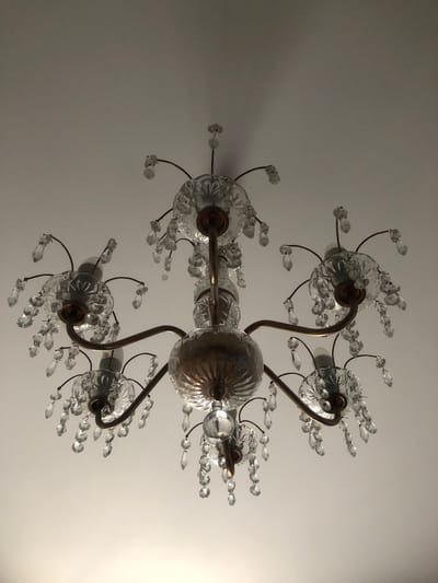 A low-angle medium shot captures a vintage crystal chandelier hanging from a white ceiling. The chandelier has a central brass body with multiple arms extending outwards, each supporting a glass fixture adorned with crystal droplets. The lighting is dim, with the chandelier bulbs emitting a soft glow that reflects off the crystals. The background is a plain white ceiling, with some light pooling at the bottom of the frame. There are no people or actions visible in the scene. The location is identified as Tutaev, Russia.